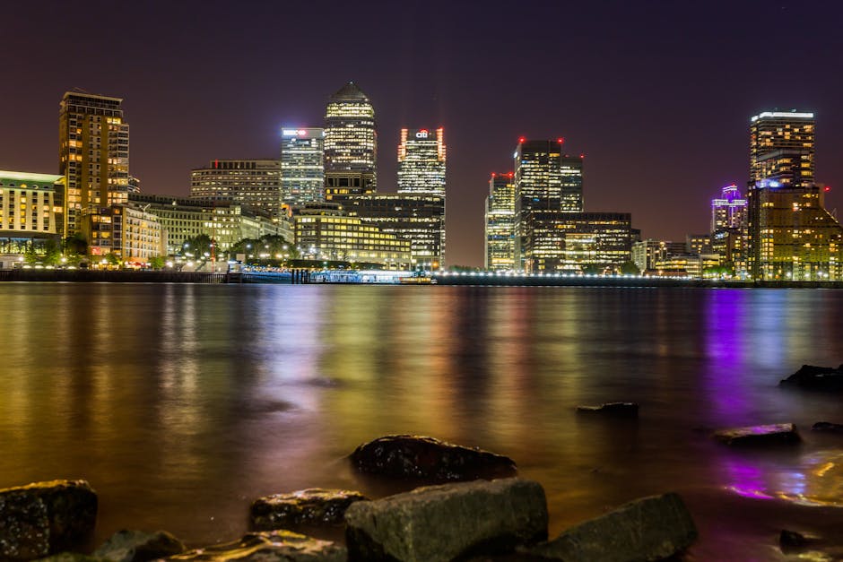 Night-time cityscape of Canary Wharf featuring a cluster of illuminated modern office buildings and skyscrapers with glass exteriors reflecting city lights. The tallest tower, centrally positioned, ha