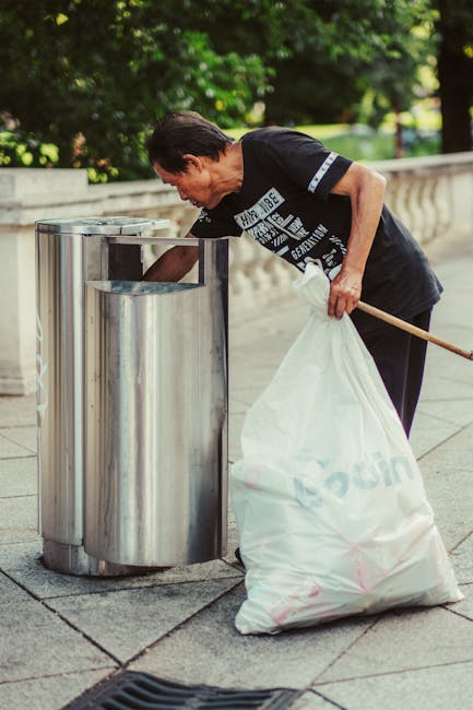 A man with dark hair wearing a black T-shirt with white graphics is seen disposing of waste into a cylindrical metal litter bin on a pavement. He is bent over, holding a large white plastic bag filled