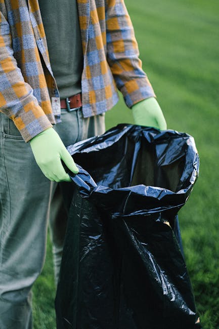 A man with dark hair wearing a black T-shirt with white graphics is seen disposing of waste into a cylindrical metal litter bin on a pavement. He is bent over, holding a large white plastic bag filled